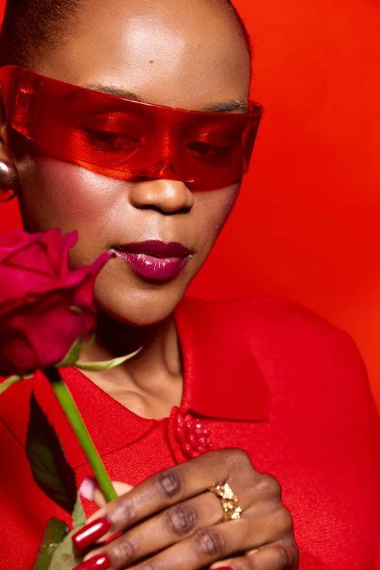 Elegant dark skinned African woman in red outfit and translucent red glasses holds a red rose close to her face. Studio portrait with vibrant red background and glossy makeup concept.