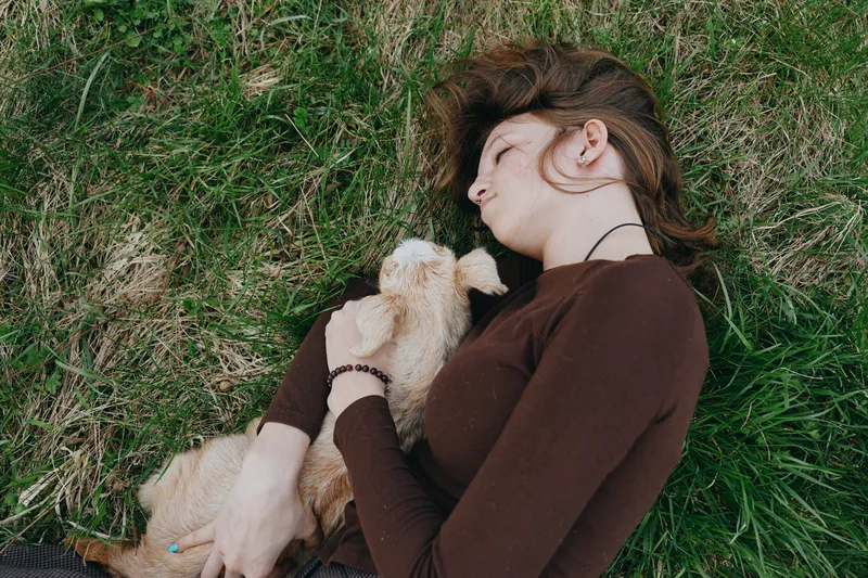Young woman with long brown hair lies on grass holding a small baby goat close to her chest in a relaxed outdoor lifestyle moment.