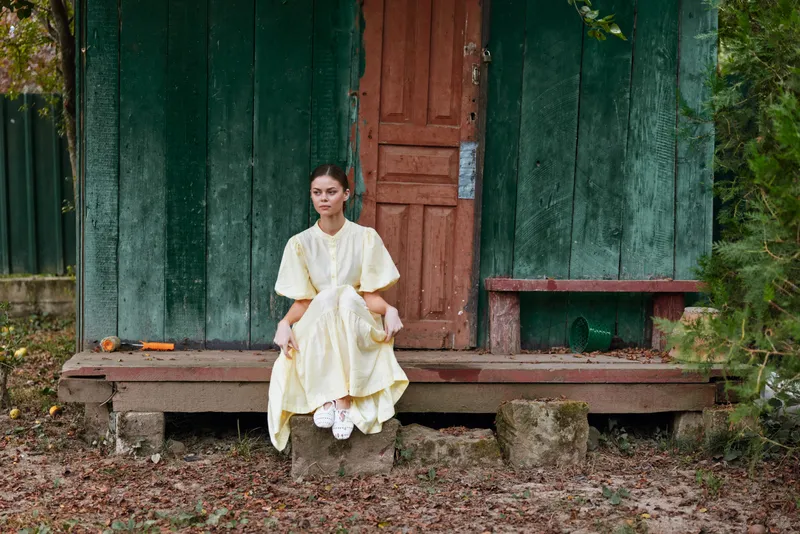 Woman in a pale yellow dress sits on the wooden steps of a green exterior building outdoors, autumn leaves scattered on the ground, creating a calm portrait and rustic scene for a natural lifestyle
