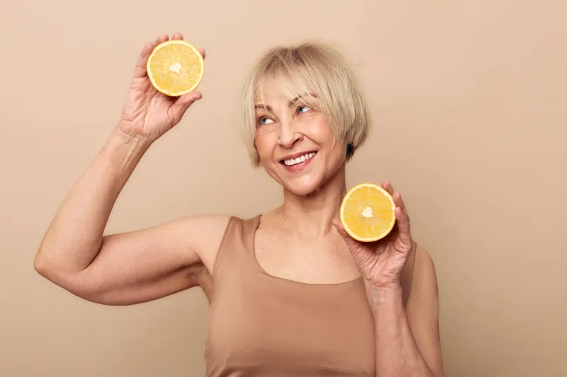 Smiling mature woman with short blonde hair holds two orange halves near face against beige background. Concept for healthy lifestyle, vitamin intake, and natural skincare.