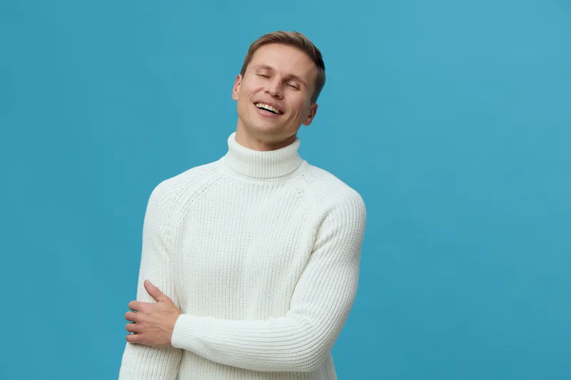 Smiling man wearing white knitted sweater against bright blue background looking happy and relaxed. Studio portrait capturing cheerful emotion and casual style for lifestyle or fashion concepts.