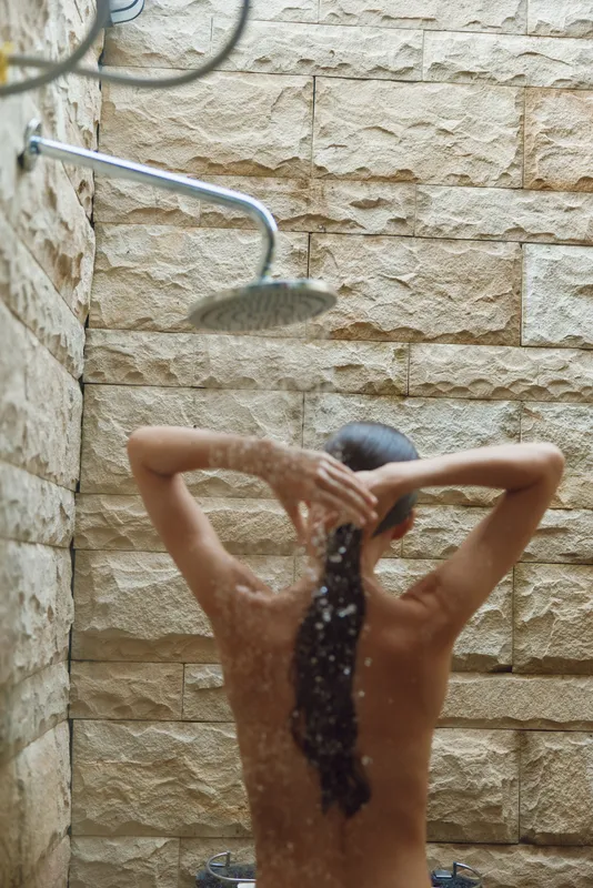 woman showering outdoors with stone wall background, long hair wet under flowing water, natural light and fresh atmosphere, relaxing moment