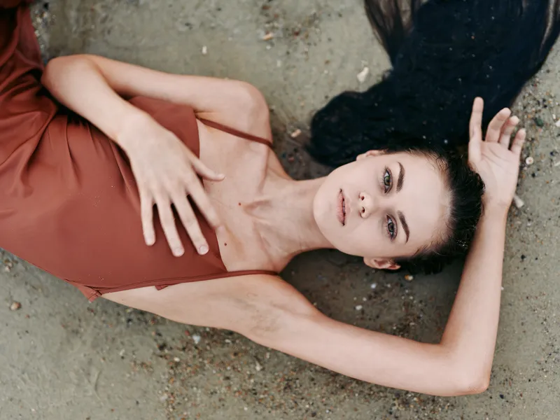 A fashion portrait of a poised woman lying on sandy ground in a warm rust dress, with soft light, subtle makeup, and relaxed elegance for a minimal, cinematic mood.
