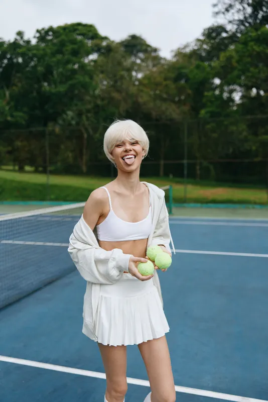 Young female tennis player with blonde hair in sporty white outfit, smiling playfully while holding tennis balls on a court surrounded by green trees