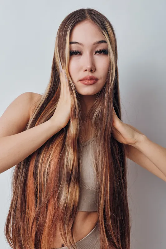 Woman with long straight hair posing with hands on face against plain background. Beauty, fashion, and hairstyle concept with natural makeup and casual outfit in studio portrait.