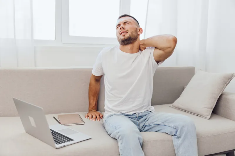 Young man experiencing neck pain while working on a laptop, sitting comfortably on a couch. Light, airy, and modern home office setting enhances the atmosphere.
