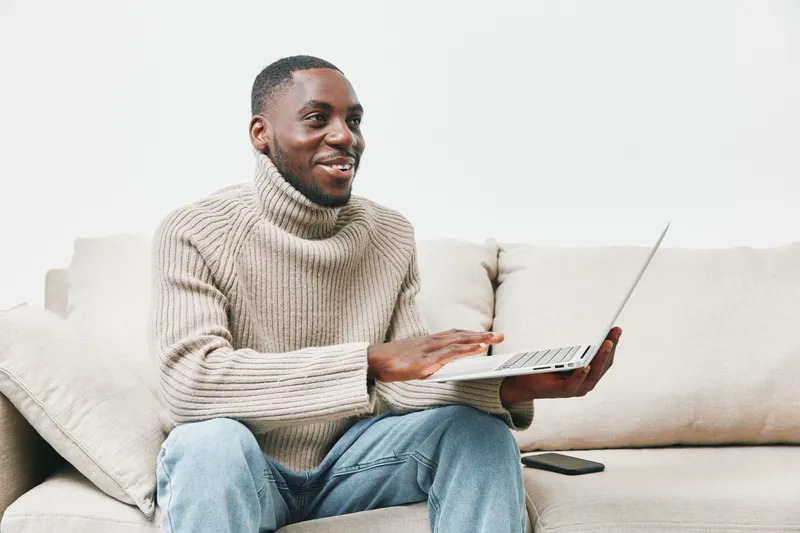 Young African man smiling while using a laptop at home, wearing a cozy sweater, sitting on a sofa, creating a relaxed and productive atmosphere