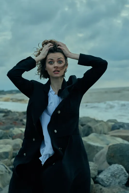 Woman in a dark coat on a rocky beach, wind-tossed hair and tense expression, stormy sky creating a moody outdoor fashion moment.