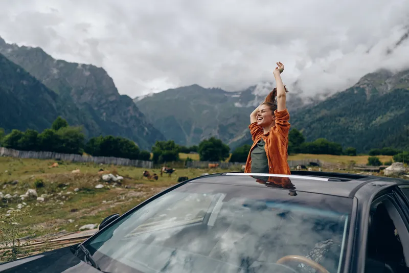 roadtrip travel woman mountains car nature inclusivity authenticity joyful woman standing through car sunroof on countryside road with mountains celebrating adventure and freedom in authentic moment