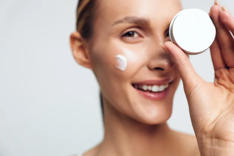 Young woman skincare, applying face cream with a small mirror, close up beauty routine on clean light background, calm morning care smile and fresh glow for skin