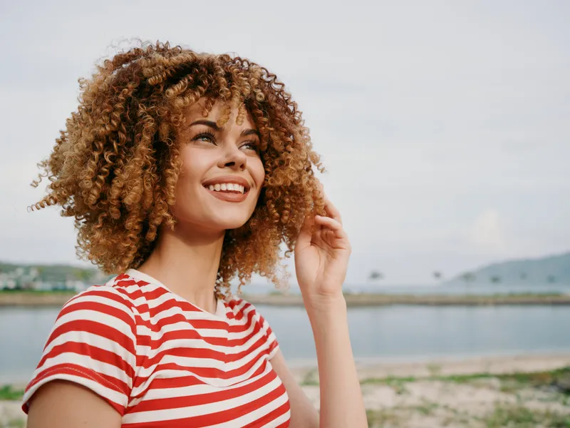 Portrait of a young woman with curly hair wearing a red striped shirt, standing outdoors by a calm body of water, smiling softly at the camera.