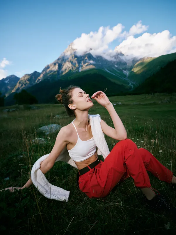 Outdoor mountains, woman in white top and red pants seated on a meadow, towel over shoulder, gazing up at blue sky with dramatic alpine backdrop