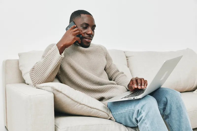 Young Black man in cozy sweater engaging in phone conversation while working on laptop at home, showcasing a warm and productive atmosphere