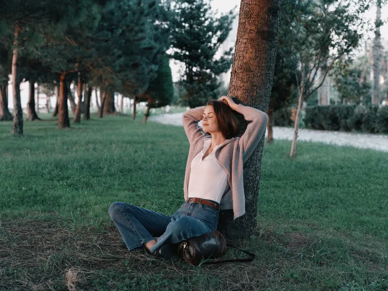 A relaxed woman sits against a tree in a park, enjoying a calm moment outdoors. She leans back, eyes closed, exuding serenity and peaceful balance.
