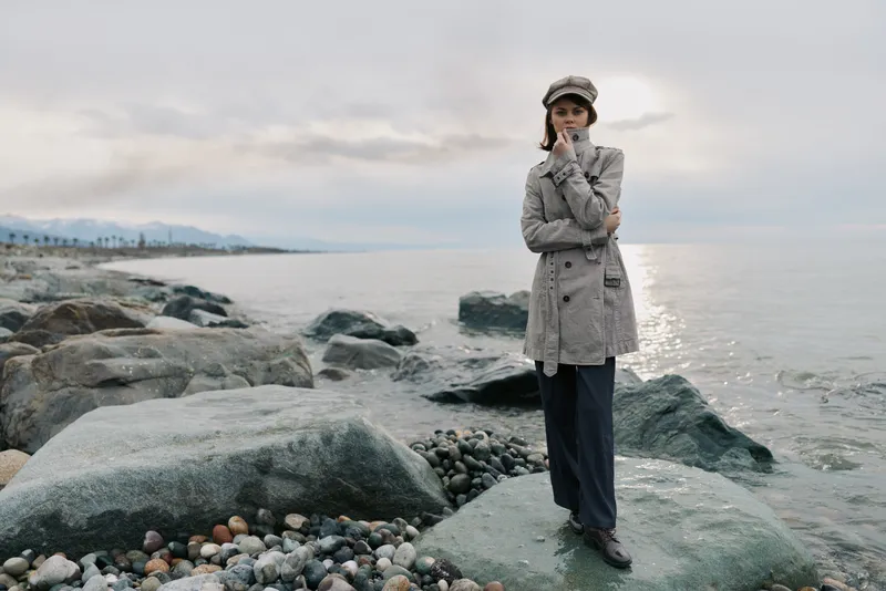 A thoughtful woman in a grey coat and hat stands on rocks by the sea under an overcast sky. The moody coastal scene conveys contemplation, calmness, and autumn fashion outdoors.