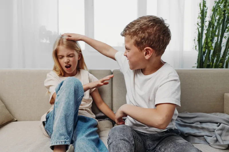 Young boy playfully teasing girl on couch in bright living room, girl reacting with surprise and discomfort, sibling interaction concept.