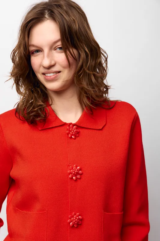 Young woman with wavy brown hair wearing a red knitted cardigan with decorative buttons smiles gently against a plain white background. Casual fashion and natural beauty concept.