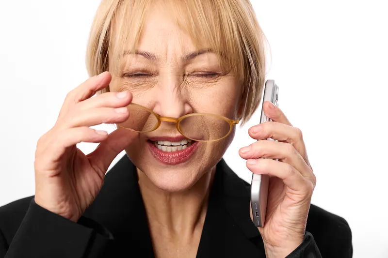 Older woman phone call glasses business communication office portrait closeup smiling, holding smartphone to ear and adjusting eyeglasses with other hand in calm studio