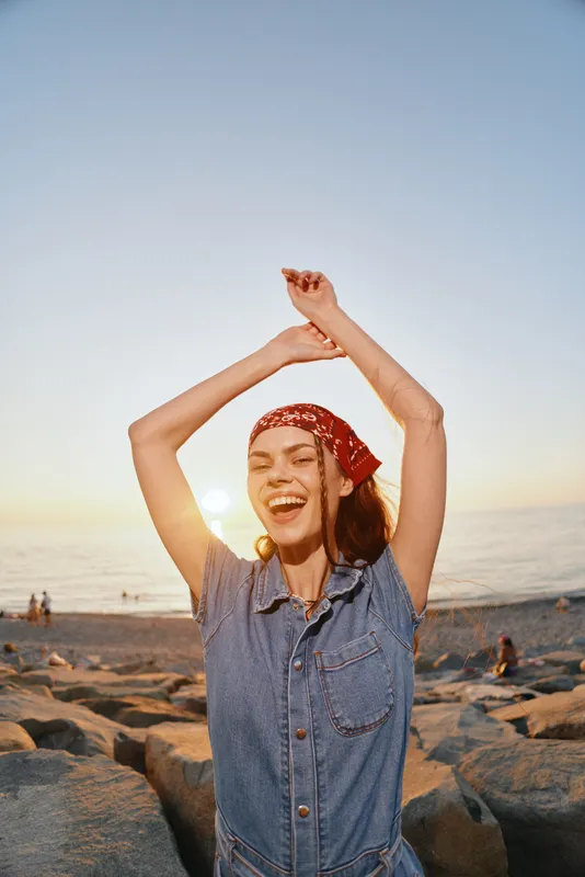 Lifestyle woman in modern boho-western denim street style smiling joyfully with arms raised at sunset beach with warm film color tones.