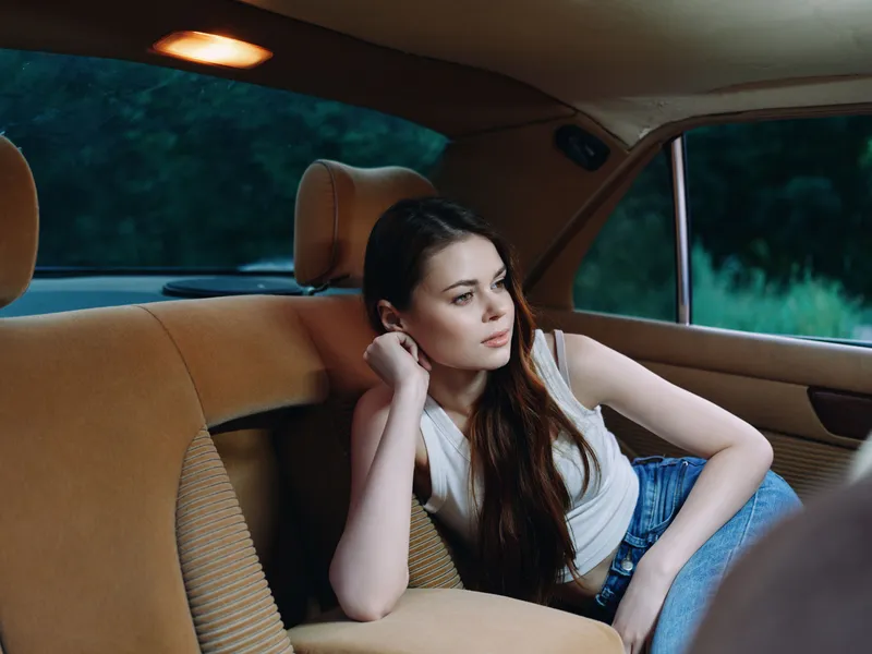Young woman with long dark hair in casual outfit, relaxed in car, showcasing contemplation and serenity, surrounded by soft beige interior in natural light.