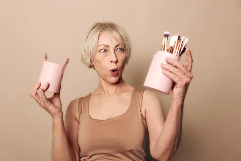 Surprised mature woman in beige tank top holds pink containers with makeup brushes, isolated on beige background. Concept for beauty tools, cosmetics, and skincare accessories.
