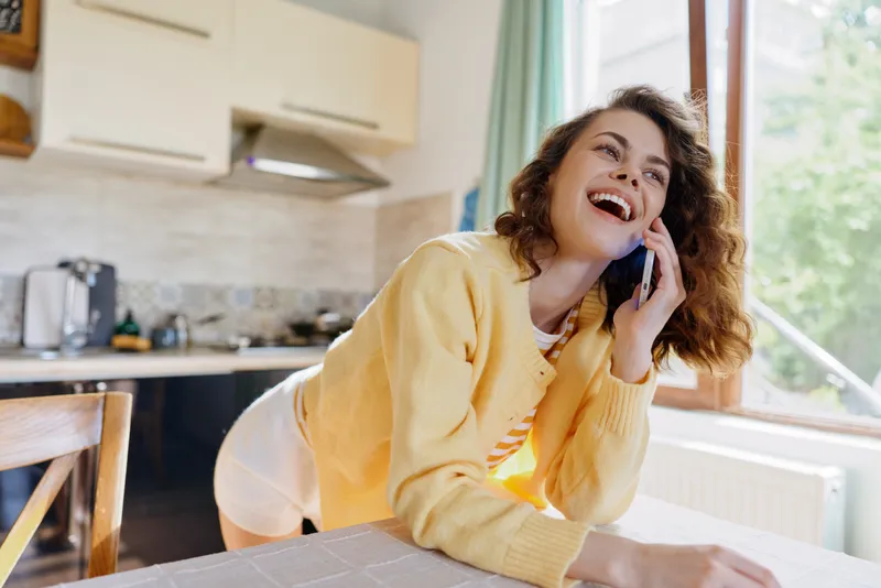 Bright kitchen scene shows a young woman with curly hair smiling while talking on a phone, leaning on a counter in a cozy home moment.
