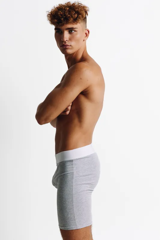 Young man with curly hair posing shirtless in gray shorts against white background, showing athletic build and confident expression in studio lighting.