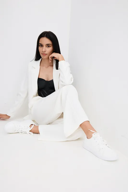 Young woman sitting on white floor wearing white pantsuit and black top, posing confidently with long black hair and white sneakers, minimalist studio background.