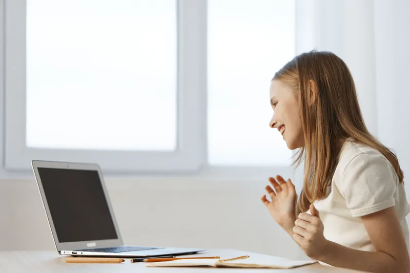 Cheerful girl using laptop for online learning while sitting at wooden desk by the window in a bright room, showcasing modern education concept