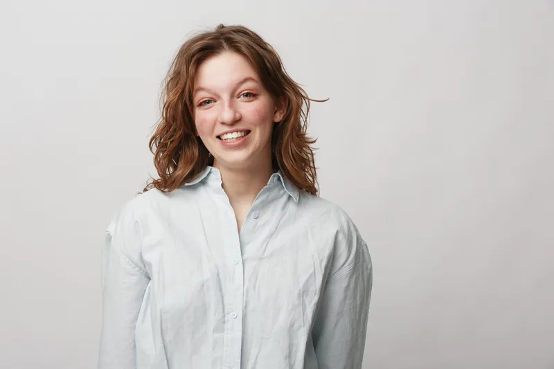 Smiling young woman with curly hair wearing a white shirt stands against a light background. Natural expression and casual style create a friendly and approachable mood.