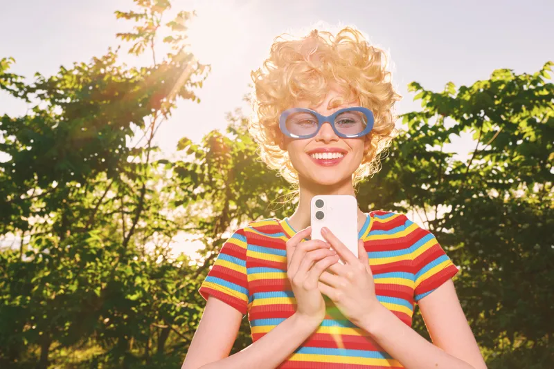 Cheerful woman outdoors, smiling in colorful striped shirt, retro glasses, sunlit moment, relaxed pose, warm breeze, vibrant greenery background, lifestyle portrait with natural vibe