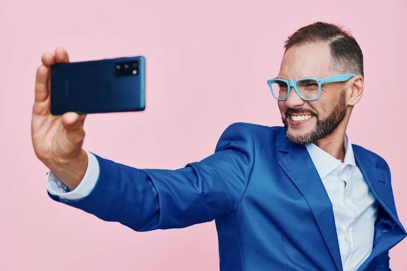 A professional man in a blue suit takes a confident selfie against a vibrant pink background, showcasing energy, focus, ambition, and a positive mood in a bold studio scene.