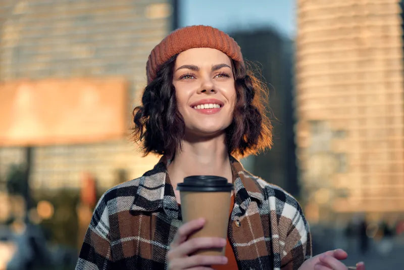 Woman with coffee in city portrait, beanie and warm smile in golden hour sunlight, candid lifestyle moment conveying authenticity, mindful living and emotional storytelling.