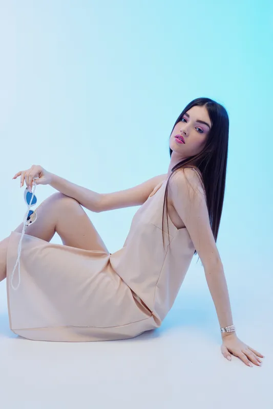Young woman in a beige dress sits on the floor holding sunglasses. She looks confident and stylish in a bright studio. Concept of woman beauty fashion and elegance.