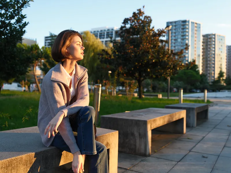 A thoughtful woman sits on a concrete bench in a modern urban park at sunset. She gazes into the distance, exuding calm, focus, and quiet confidence in a serene, cinematic atmosphere.