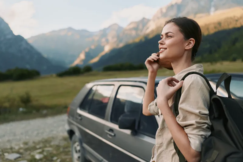 Young woman with a backpack stands near a car on a mountain road enjoying the fresh air and scenic view with a relaxed and happy expression.