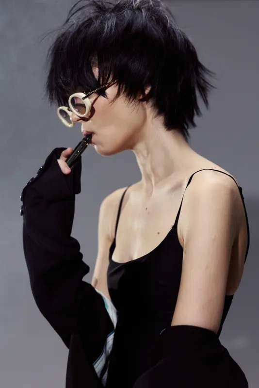 Stylish woman in black outfit with white glasses posing against gray background, modern fashion, edgy hairstyle