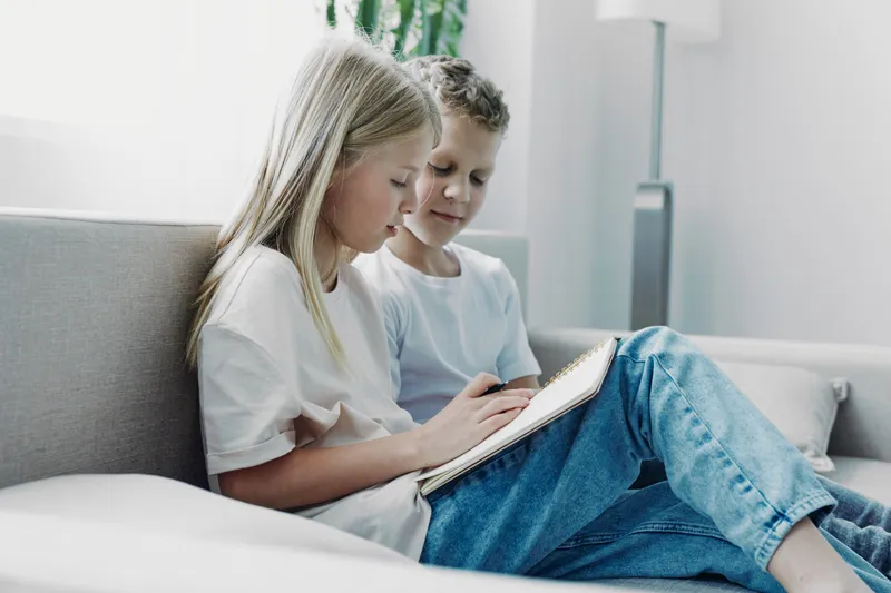 Two children sitting on a couch reading a book together in a bright living room. Casual clothes and relaxed atmosphere emphasize learning and sibling bonding at home.