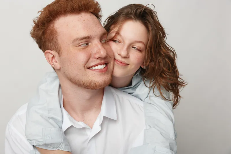 Happy young couple with red hair and casual clothes smiling and embracing each other on white background. Concept of love, friendship, relationship, and positive emotions.