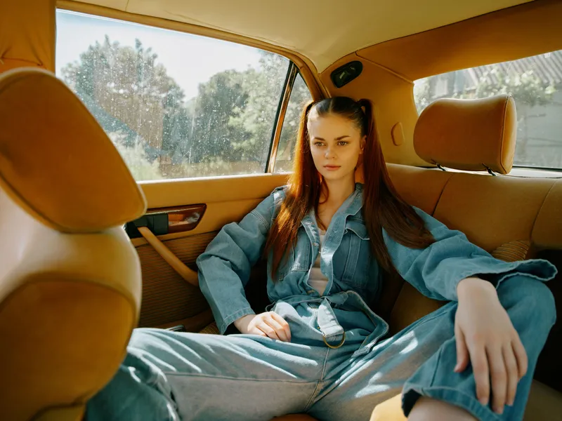 A young woman in a denim outfit relaxing in a car, showcasing casual style and confidence in a sunny setting, with a natural outdoor background.