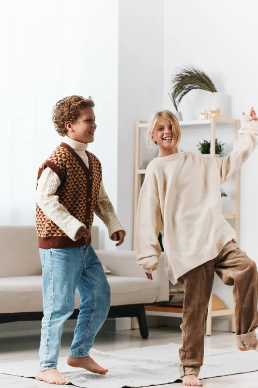 Two cheerful children dance indoors wearing cozy knit sweaters and comfy jeans, their bright smiles and energetic steps highlighting a spontaneous moment of friendship and joy.
