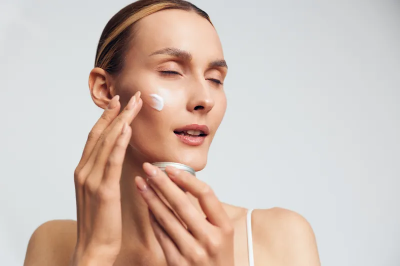 Woman applying face cream on her cheek, close up beauty skincare portrait, eyes closed, soft studio light on light background, conveying wellness and calm self confidence