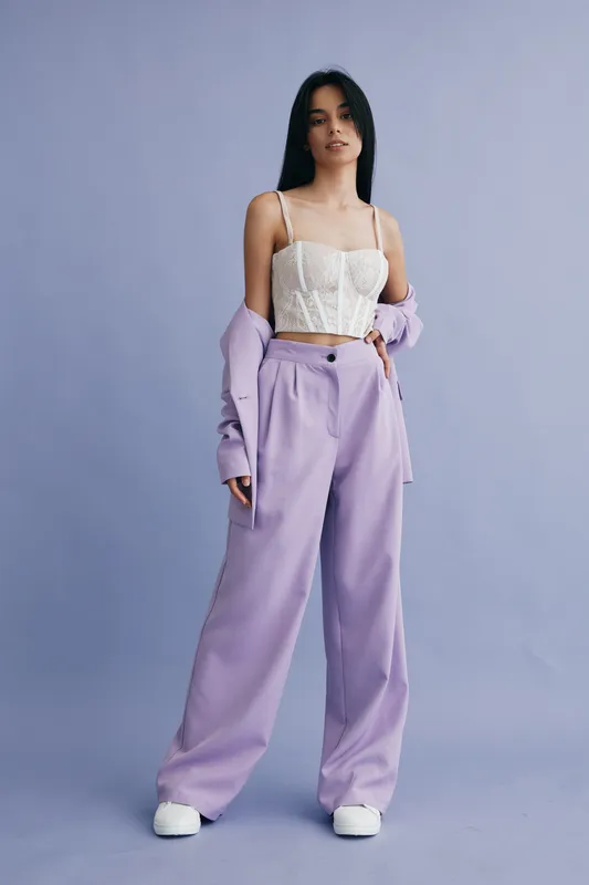 young woman wearing white corset top and oversized lilac pants stands against plain purple background, stylish casual outfit, modern fashion look, confident pose