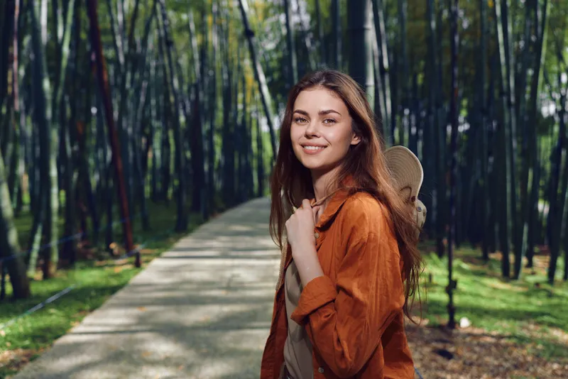 Woman with backpack smiling on a park path, forest nature portrait of a young traveler enjoying sunlight and green foliage. Casual jacket, walking tour and peaceful outdoor leisure moment.