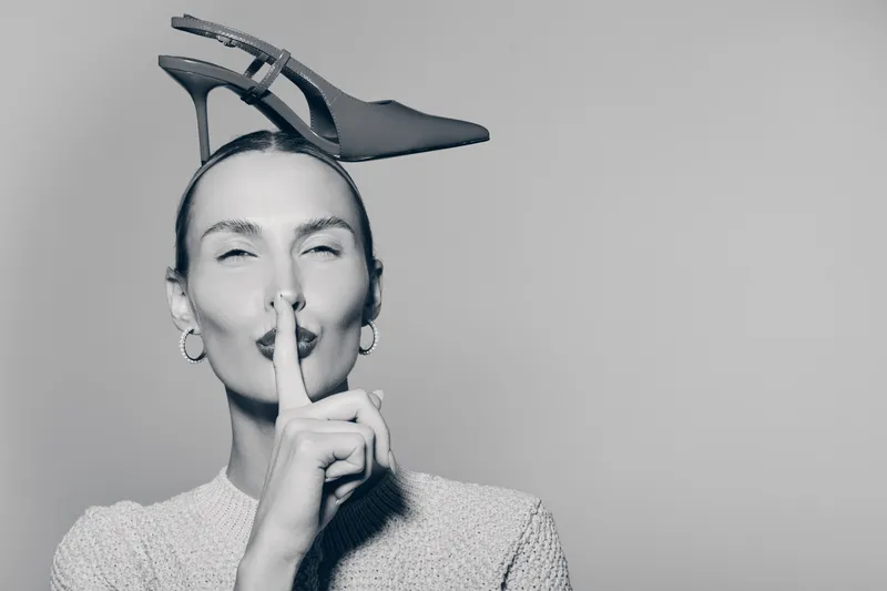 Young white woman with pale skin holds finger to lips in shushing gesture while balancing high heel shoe on head in black and white portrait.