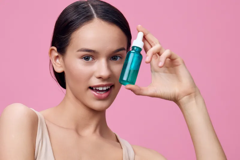 Young woman showcasing a green serum bottle against a pastel pink background, promoting skincare and beauty concepts in a fresh, vibrant atmosphere