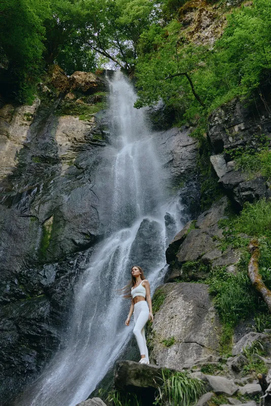 woman standing near tall waterfall surrounded by lush green forest and rocky cliffs in bright outdoor nature setting during daytime