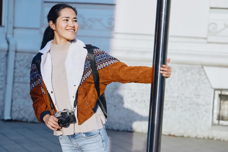 Woman with a camera and warm patterned jacket stands outdoors beside a street lamp, smiling, ready for candid travel photography in an urban setting.