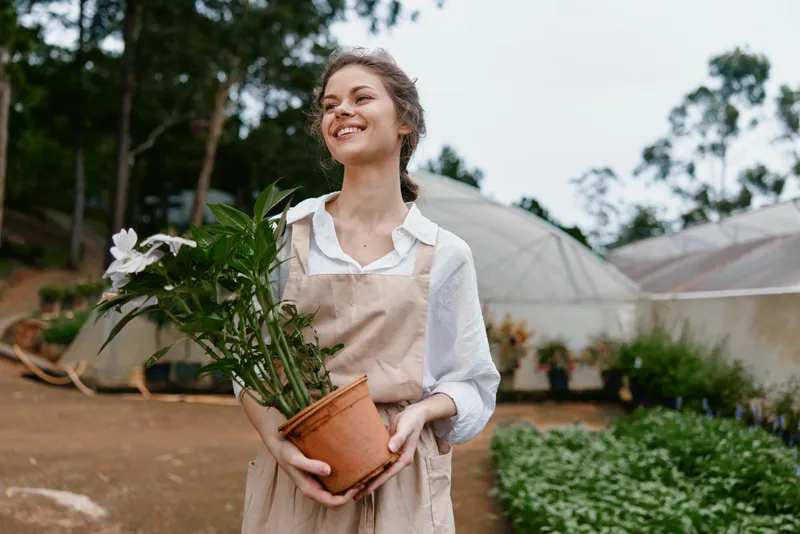 Young woman smiling while holding potted plant in garden, wearing casual clothing, surrounded by greenery and greenhouse