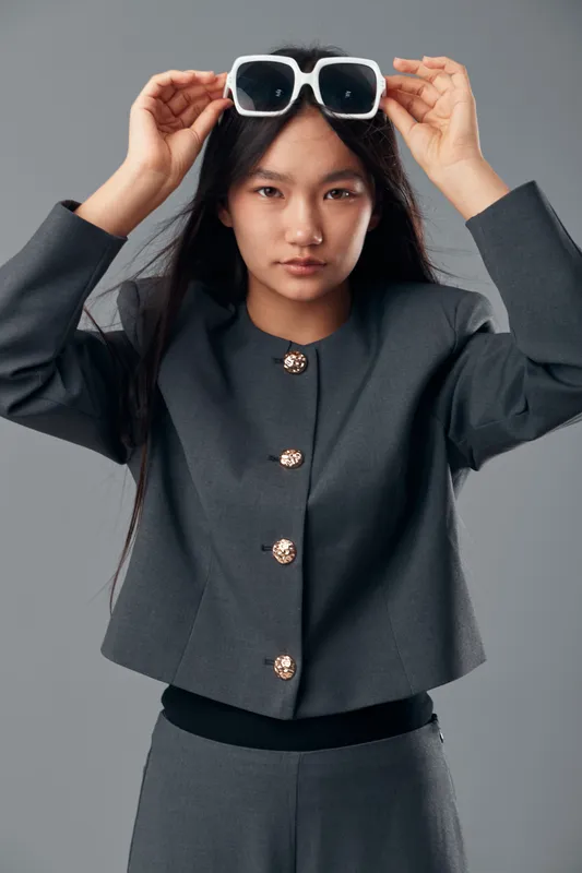 Young woman with long black hair wearing a gray suit adjusting white sunglasses on head against gray background, studio portrait with fashionable modern outfit and serious expression.